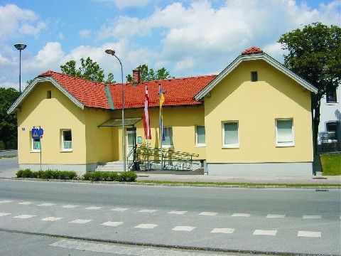 Ein gelbes Gebäude mit rotem Dach und mehreren Fenstern steht an der Straße. Zwei Fahnen sind auf Stangen vor dem Gebäude gehisst. Eine Straßenlaterne befindet sich auf der linken Seite des Gebäudes.