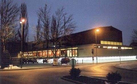 Ein großes Gebäude mit Glasfenstern und Lichtern in der Nacht, umgeben von Bäumen und Straßenlaternen, mit geparkten Autos und einem Schild vorne.