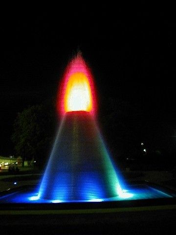 Ein Brunnen mit kegelförmiger Form strahlt nachts mehrfarbiges Wasser aus. Die Wasserfarben umfassen Blau, Grün, Gelb und Rot. Das Wasser schießt nach oben und erzeugt einen Regenbogeneffekt.