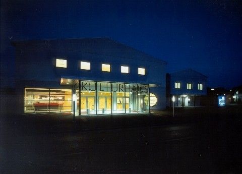 Die Außenseite eines modernen Gebäudes, beschriftet mit 'Kulturhaus', ist bei Nacht beleuchtet. Es gibt große Glasfenster, einen prominenten Eingang und mehrere beleuchtete Fenster.