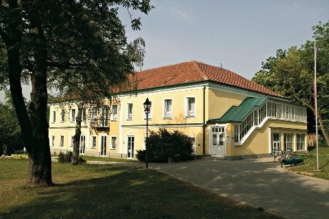 Ein gelbes Gebäude mit rotem Dach und grüner Einfassung, in einem Park mit Bäumen und Rasen gelegen, hat mehrere Fenster und eine Treppe, die zum Eingang führt.