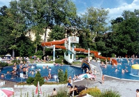 Ein großer Außen-Wasserpark mit mehreren Pools, Rutschen und schwimmenden Menschen. Es gibt Bäume und Sonnenschirme rund um den Park.