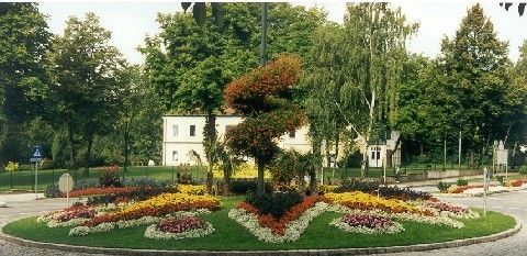 Ein runder Garten mit bunten Blumen und einem weißen Gebäude im Hintergrund. Bäume umgeben den Garten, und ein blaues Schild befindet sich links.