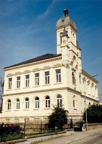 Ein großes weißes Gebäude mit Rundbogenfenstern und einem Glockenturm steht an einer Straße mit Zaun und Straßenlaterne.