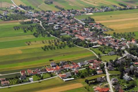 Luftaufnahme eines Dorfes mit mehreren Häusern, umgeben von grünen Feldern und Bäumen.