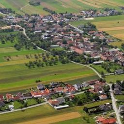 Luftaufnahme eines Dorfes mit mehreren Häusern, umgeben von grünen Feldern und Bäumen.