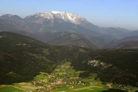 Eine Berglandschaft mit schneebedeckten Gipfeln, einem Tal darunter und einem Dorf, das zwischen grünen Feldern und dichten Wäldern eingebettet ist.