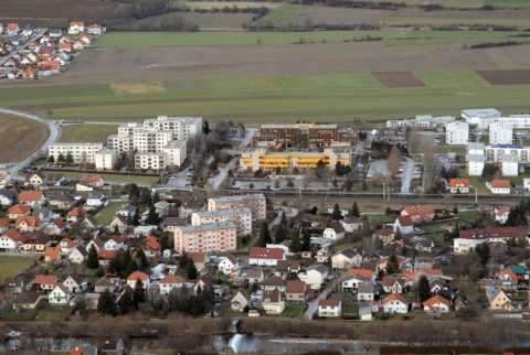 Luftaufnahme einer Stadt mit verschiedenen Gebäuden, umgeben von grünen Feldern und Bäumen, und einer Eisenbahnlinie in der Mitte.
