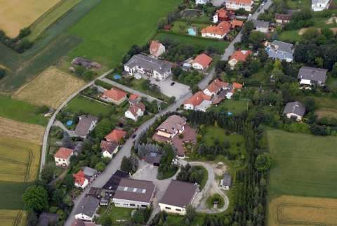 Luftaufnahme eines kleinen Dorfes mit mehreren Häusern und Feldern. Einige Häuser haben rote Dächer, andere graue Dächer. Das Dorf ist von grünen Bäumen und Büschen umgeben.