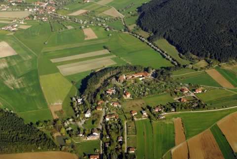 Luftaufnahme eines ländlichen Gebiets mit Häusern, Feldern und einem kleinen Wald. Das Gebiet ist von üppigen grünen Feldern und Bäumen umgeben.