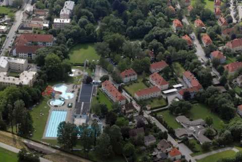 Luftaufnahme einer Stadt mit einem Schwimmbad und verschiedenen Gebäuden mit roten Dächern. Bäume und Grünflächen umgeben das Gebiet.