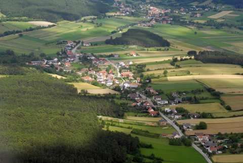 Luftaufnahme einer kleinen Stadt mit Häusern und grünen Feldern, umgeben von Bäumen und einer Straße. Die Stadt liegt in einem Tal mit klarem Himmel darüber.