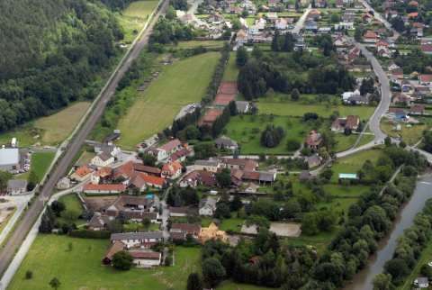 Luftaufnahme eines ländlichen Dorfes mit dachbedeckten Häusern, umgeben von grünen Feldern, Bäumen und einem Fluss. Das Dorf liegt zwischen einer Hauptstraße und einer kleineren Straße.