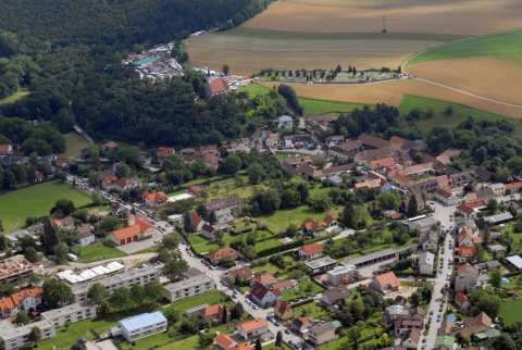 Luftaufnahme einer Stadt mit zahlreichen Gebäuden, Straßen und üppiger Grünfläche. Bauernhäuser mit roten Dächern sind über die Stadt verstreut.