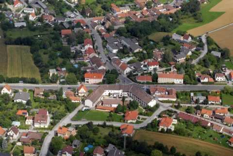 Luftaufnahme eines kleinen Dorfes mit vielen Häusern, einige mit roten Dächern, umgeben von grünen Feldern und Bäumen.