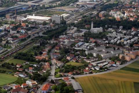 Luftaufnahme einer Stadt mit vielen Gebäuden, Straßen und Grünflächen. Im Zentrum ist ein hohes Gebäude zu sehen. Die Stadt ist von Feldern und Bäumen umgeben.