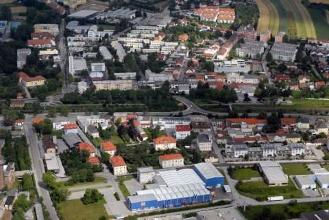Luftaufnahme eines städtischen Gebiets mit vielen Gebäuden, Straßen und Grünflächen. Bäume sind verstreut.