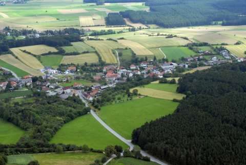 Eine Luftaufnahme einer ländlichen Landschaft mit vielen Häusern, grünen Feldern und dichten Wäldern. Das Dorf liegt in einem Tal mit Hügeln im Hintergrund.