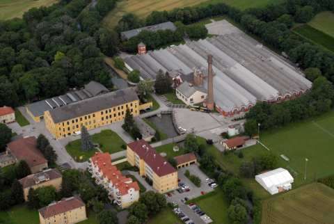Eine Luftaufnahme eines großen Industriekomplexes mit mehreren Gebäuden, einem Parkplatz und einem hohen Schornstein. Grüne Felder und Bäume umgeben das Gebiet.