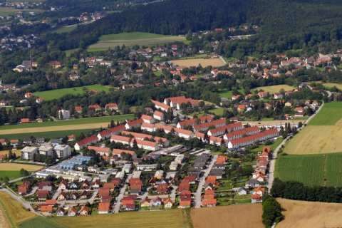 Luftaufnahme einer Stadt mit vielen Gebäuden und roten Dächern, umgeben von Feldern und Bäumen.