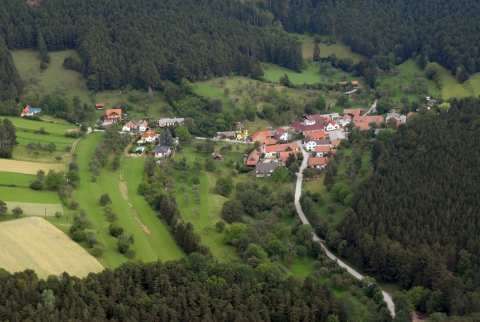 Luftaufnahme eines kleinen Dorfes in einem bewaldeten Tal mit verstreuten Häusern und grünen Feldern.