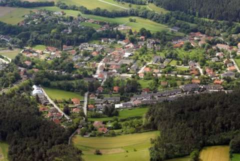 Luftaufnahme eines kleinen Dorfes mit vielen Häusern, umgeben von grünen Feldern und Bäumen, in einer ländlichen Gegend.