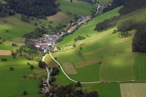 Luftaufnahme eines kleinen Dorfes, umgeben von grünen Feldern, Bäumen und einer gewundenen Straße. Das Dorf liegt in einem Tal, mit einem Gebirgszug im Hintergrund.