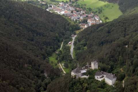 Eine Burg liegt eingebettet in einem üppigen Tal, umgeben von dichtem Wald, mit einem kleinen Dorf und Feldern in der Ferne sichtbar.