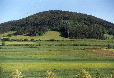 Ein gelber Heißluftballon schwebt über einem bewaldeten Hügel und bietet einen Blick auf eine weitläufige, grüne Landschaft mit kultivierten Feldern und einem klaren blauen Himmel.