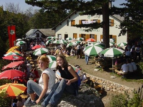 Zwei Mädchen sitzen auf einem Felsen in einem Park mit vielen Tischen und Schirmen, Menschen in der Nähe und einem weißen Gebäude im Hintergrund.