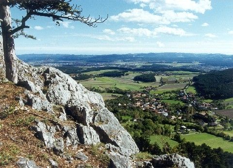 Ein Panoramablick von einem felsigen Felsvorsprung zeigt ein üppiges grünes Tal darunter mit mehreren Gebäuden und Bäumen. In der Ferne sind Berge unter einem blauen Himmel mit verstreuten Wolken zu sehen.
