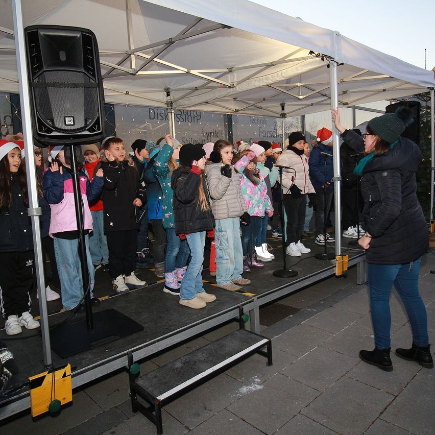 Eine Gruppe von Kindern in Winterkleidung steht auf einer Bühne unter einem Zelt und singt, während eine Frau ein Mikrofon hält.