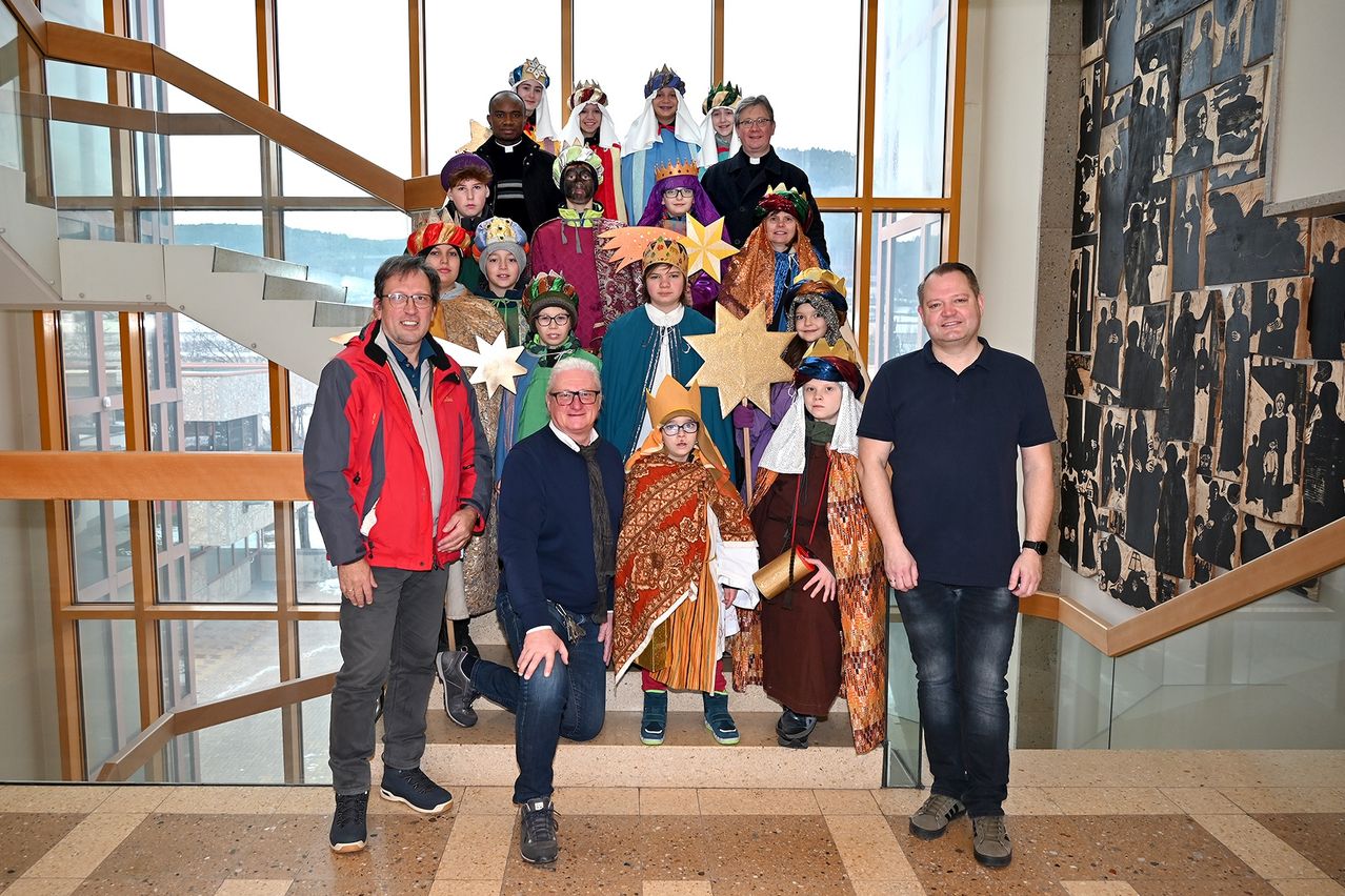 Eine Gruppe von Menschen in Weihnachtskostümen, darunter Könige und Engel, posiert mit zwei Männern, einem knienden und einem stehenden, vor einem großen Fenster.