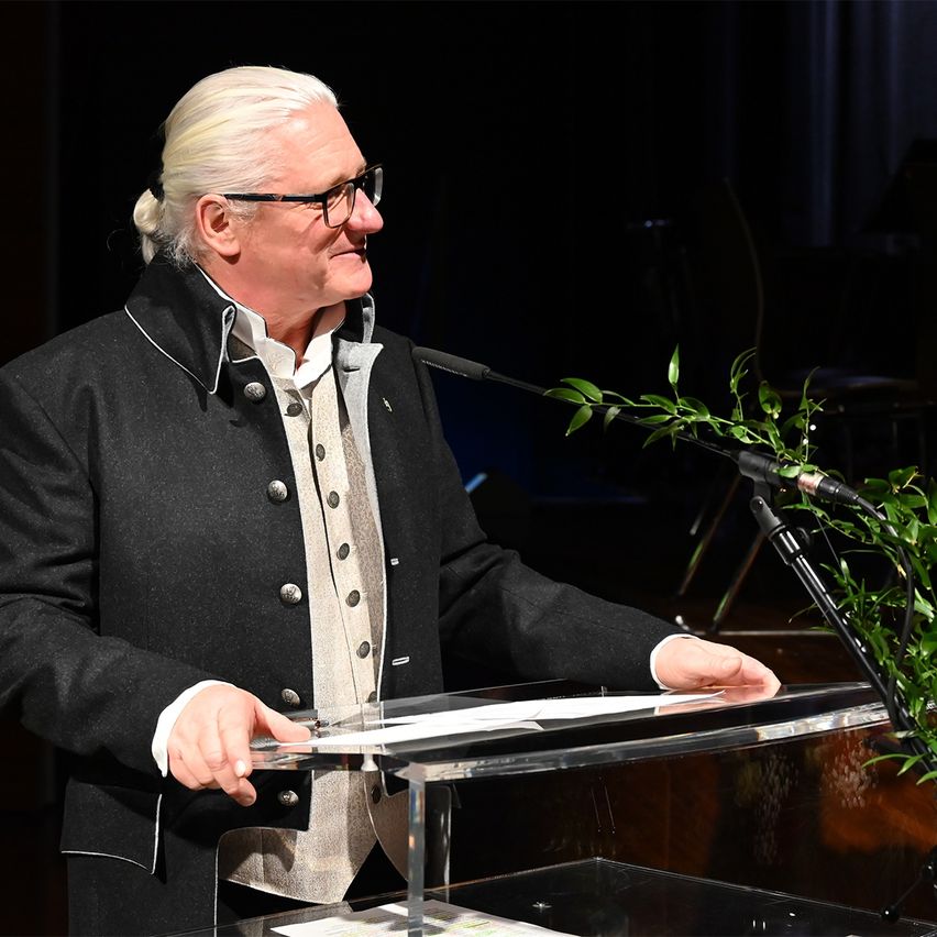 Ein älterer Mann in einem schwarzen Mantel mit einem weißen Hemd darunter steht an einem Rednerpult und hält ein Stück Papier in der Hand. Er scheint eine Rede zu halten.