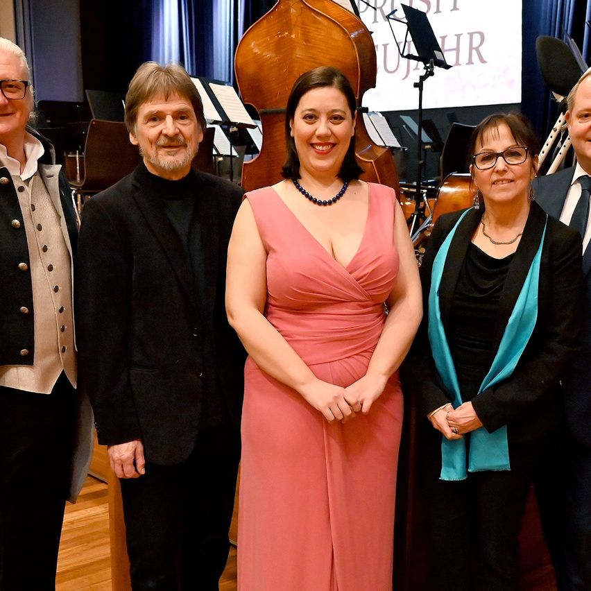 Eine Gruppe von Menschen in formeller Kleidung steht zusammen mit Musikinstrumenten im Hintergrund. Eine Frau in einem rosa Kleid lächelt und posiert für das Foto.