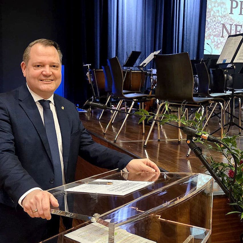 Ein Mann steht lächelnd an einem Podium in einem Konferenzraum. Papiere und ein Stift liegen auf dem Podium, und Musikinstrumente sind hinter ihm.