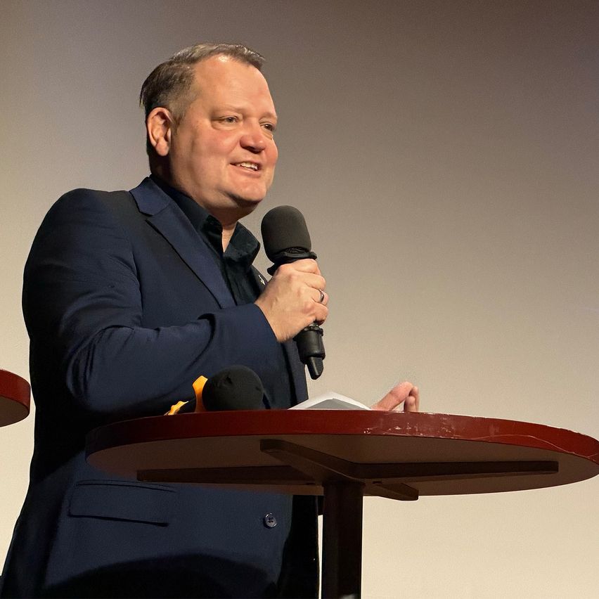 Ein Mann in einem dunkelblauen Anzug spricht in ein Mikrofon an einem Podium mit rotem Tisch. Er lächelt und hält ein Stück Papier.