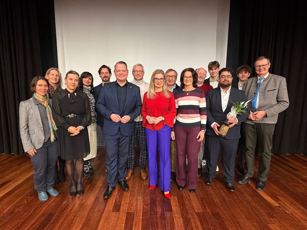 Eine Gruppe von Menschen, wahrscheinlich ein Team, posiert für ein Foto in einem Raum mit Holzboden. Eine Frau in Rot steht in der Mitte, während andere formelle Kleidung tragen. Ein Mann hält eine Topfpflanze.