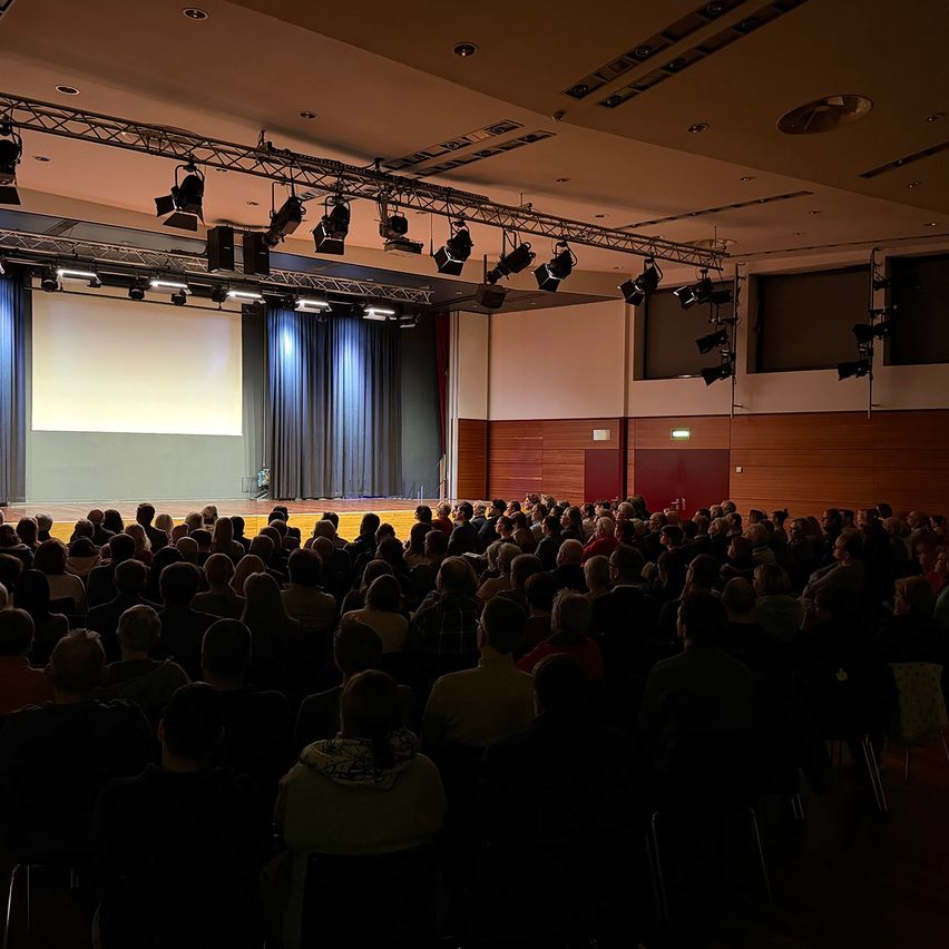 Ein Auditorium mit Bühne und Leinwand, gefüllt mit einer Menschenmenge, die auf Stühlen sitzt. Scheinwerfer hängen von der Decke.