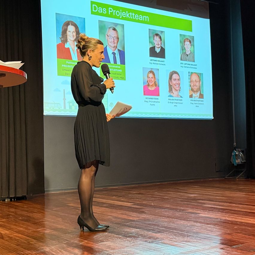 Eine Frau steht auf der Bühne und spricht in ein Mikrofon. Ein großer Bildschirm hinter ihr zeigt eine Präsentation mit dem Titel 'Das Projektteam' und Fotos mehrerer Teammitglieder.