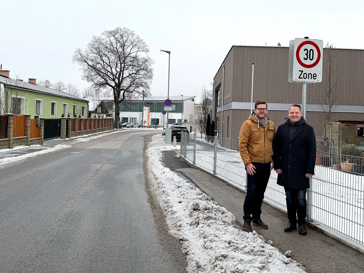 Zwei Männer stehen auf dem Bürgersteig vor einem Gebäude mit einem '30 Zone'-Schild. Der Boden ist schneebedeckt, und ein laubloser Baum befindet sich in der Nähe. Hinter ihnen steht ein Metallzaun, und eine Straße mit einem Gebäude und geparkten Autos ist sichtbar.