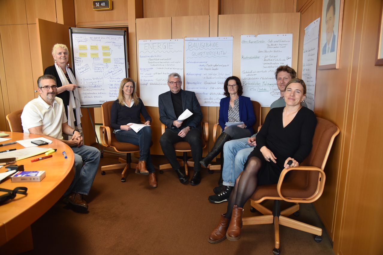 Eine Gruppe von Menschen sitzt in Stühlen in einem Raum. Sie lächeln und scheinen für ein Foto zu posieren. Ein Whiteboard mit einigen Schriftzügen steht links.