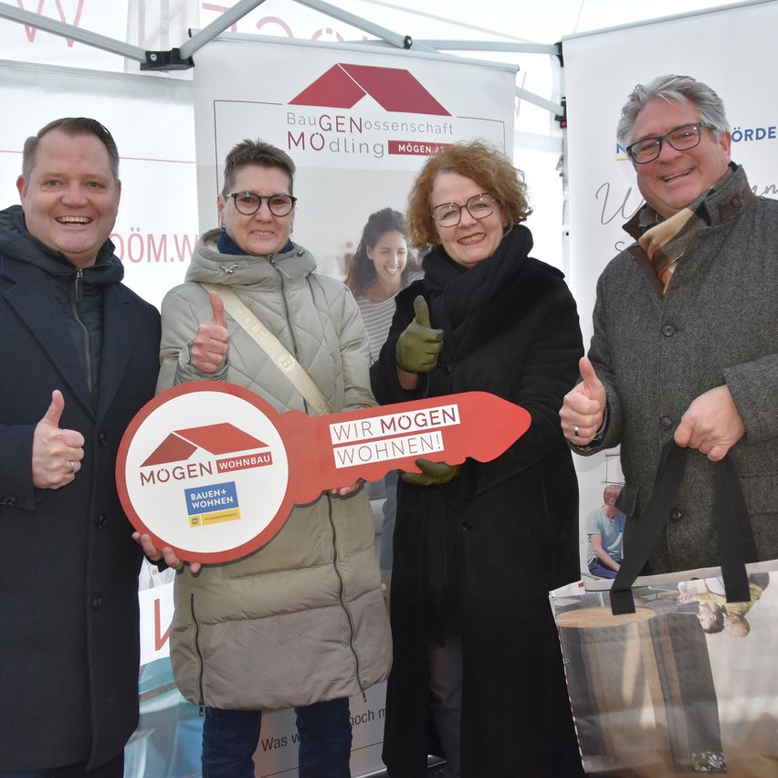Vier Personen stehen in einem Stand mit einem Banner. Sie halten ein Schild mit den Worten 'Wir Mogen Wohnen!' und lächeln für die Kamera. Dahinter ist ein großes Schild mit einem Bild einer Frau.