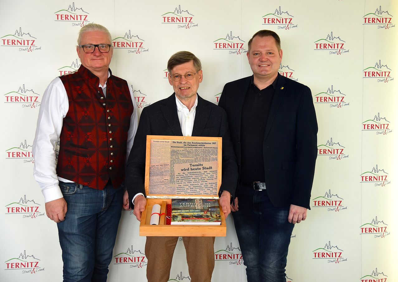 Drei Männer stehen zusammen, einer hält ein Buch in einer Holzkiste und lächelt. Sie stehen vor einer Wand mit 'TERNITZ' Logos.