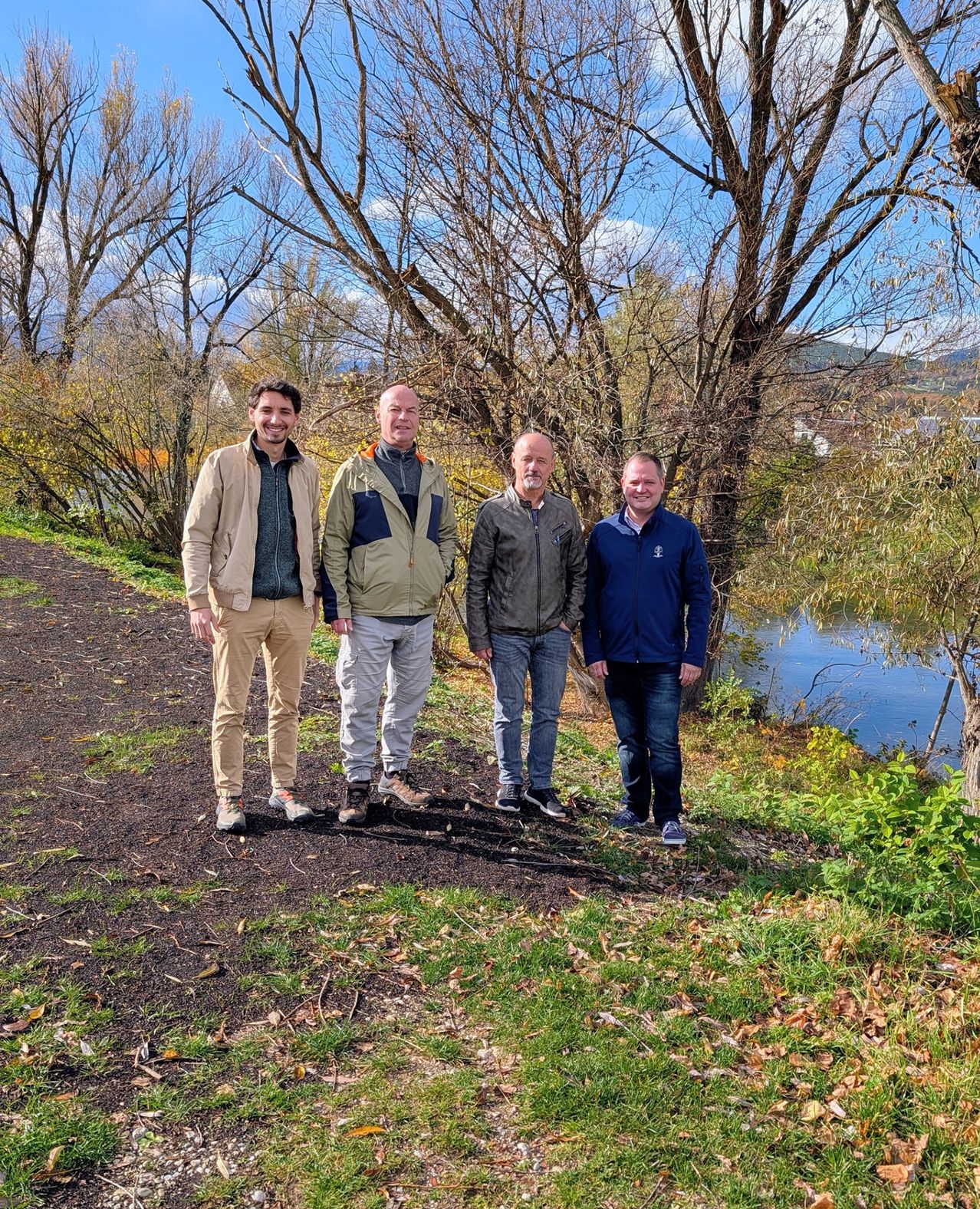Vier Männer stehen auf einem Pfad neben einem Fluss und lächeln für ein Foto. Sie sind von Bäumen und einem klaren blauen Himmel umgeben.