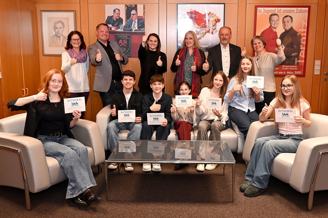 Eine Gruppe von Menschen, Erwachsene und Kinder, posiert für ein Foto in einem Raum mit einem Glastisch und Bilderrahmen an der Wand. Alle halten ein Schild mit dem Wort 'Gutschein' darauf.