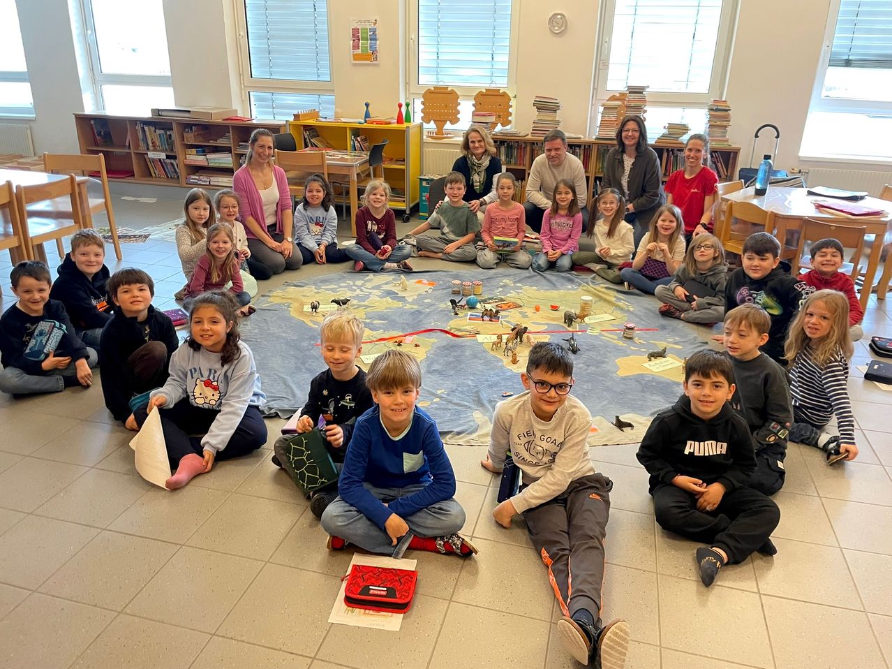 Eine Gruppe von Kindern und Erwachsenen in einem Klassenzimmer, die um eine Karte mit Spielzeug herumsitzen.