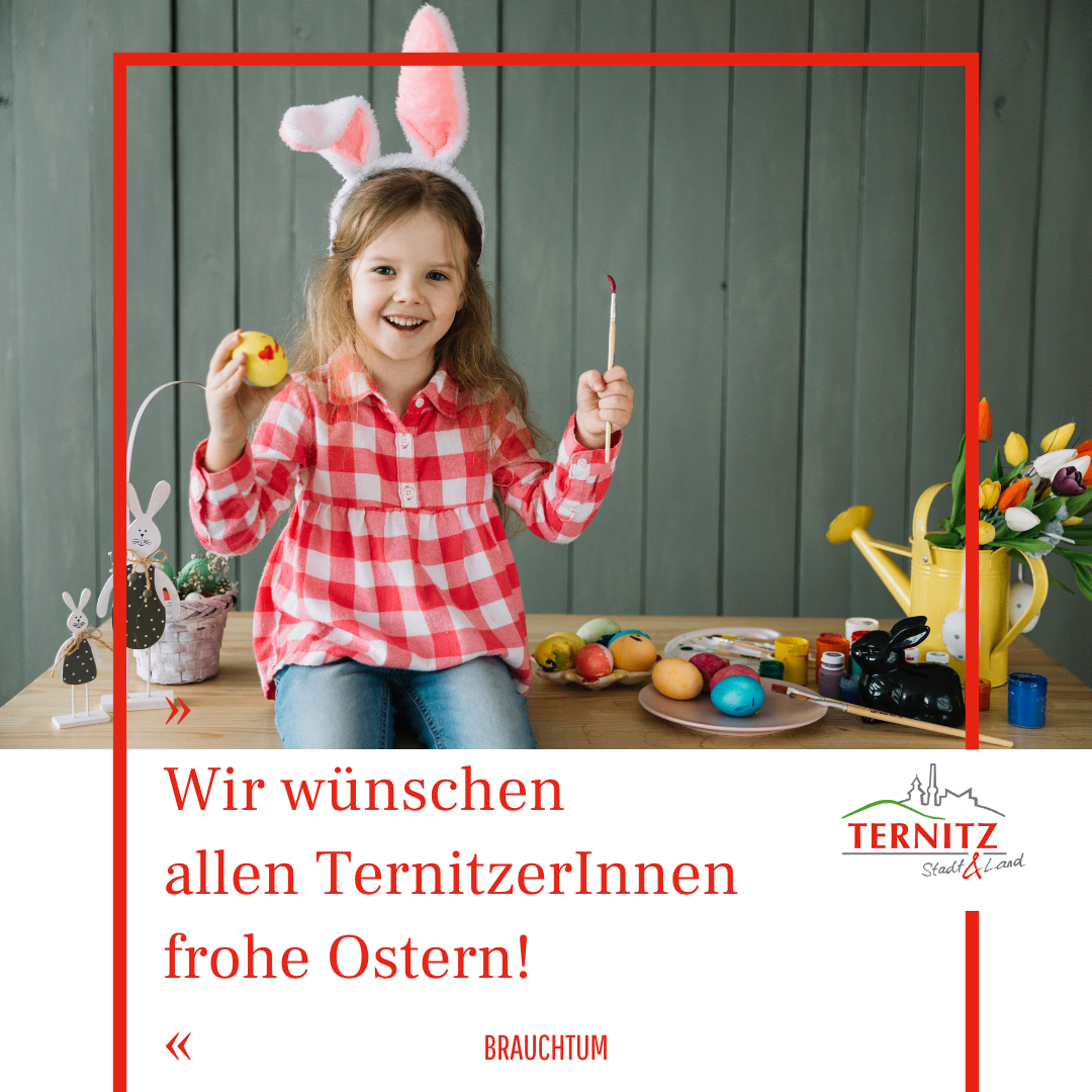 Ein kleines Mädchen mit Hasenohren hält ein bemaltes Osterei in der Hand, umgeben von anderen Eiern und einer Hasenfigur. Hinter ihr steht ein Tisch mit einem Korb und Blumen. Der Text lautet 'Wir wünschen allen TernitzerInnen frohe Ostern!'