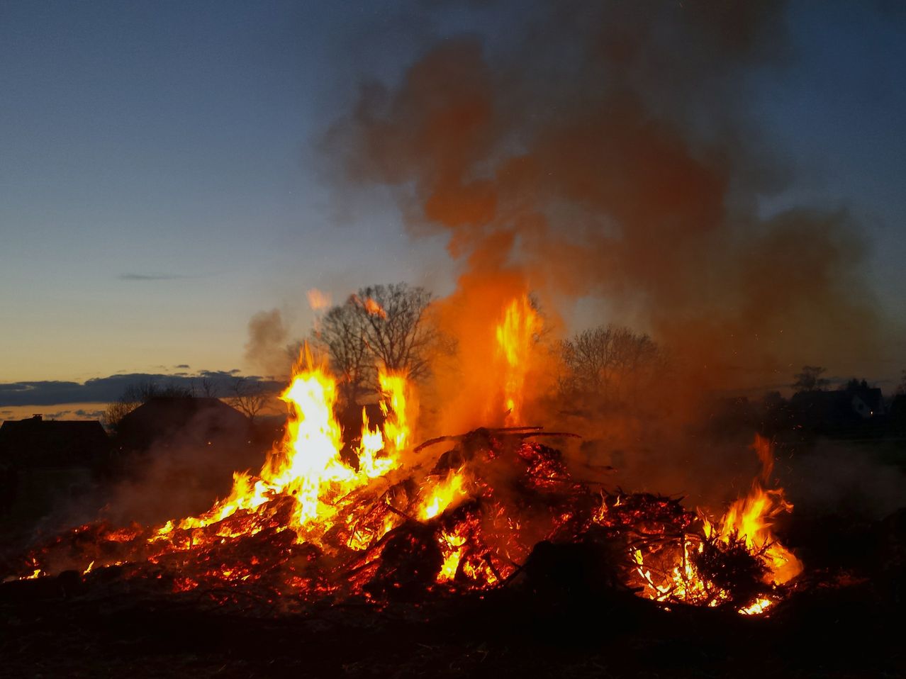 Ein großes Lagerfeuer brennt intensiv in der Dämmerung und wirft ein oranges Licht auf die Silhouette der kahlen Bäume. Das Feuer sendet dicken Rauch aus, der in den sich verdunkelnden Himmel aufsteigt.
