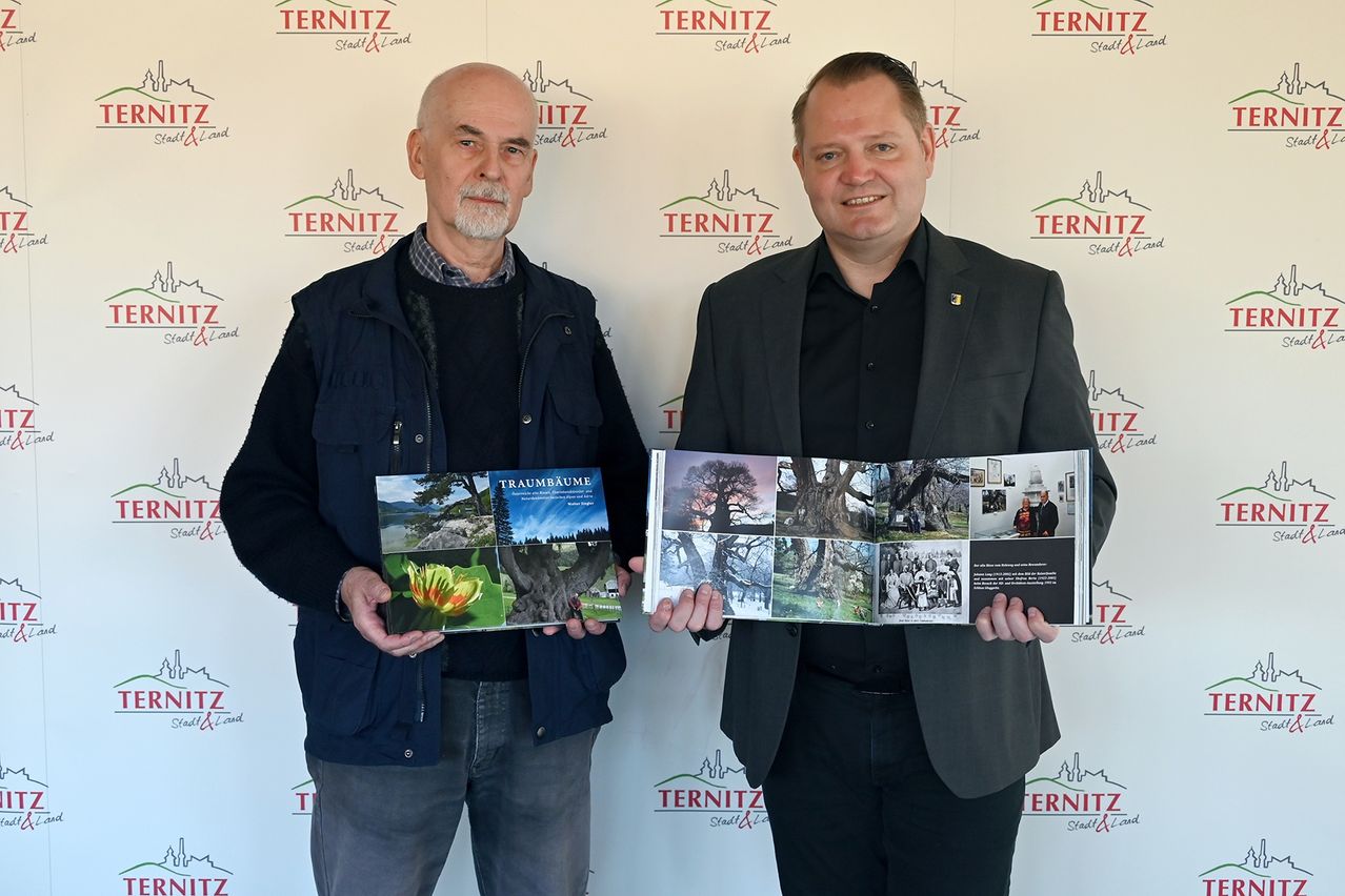 Zwei Männer stehen vor einem Hintergrund mit Logos und Text. Der Mann auf der linken Seite hält ein Buch mit dem Titel Traumbaume. Der Mann auf der rechten Seite hält ein größeres Buch mit vielen Fotos.